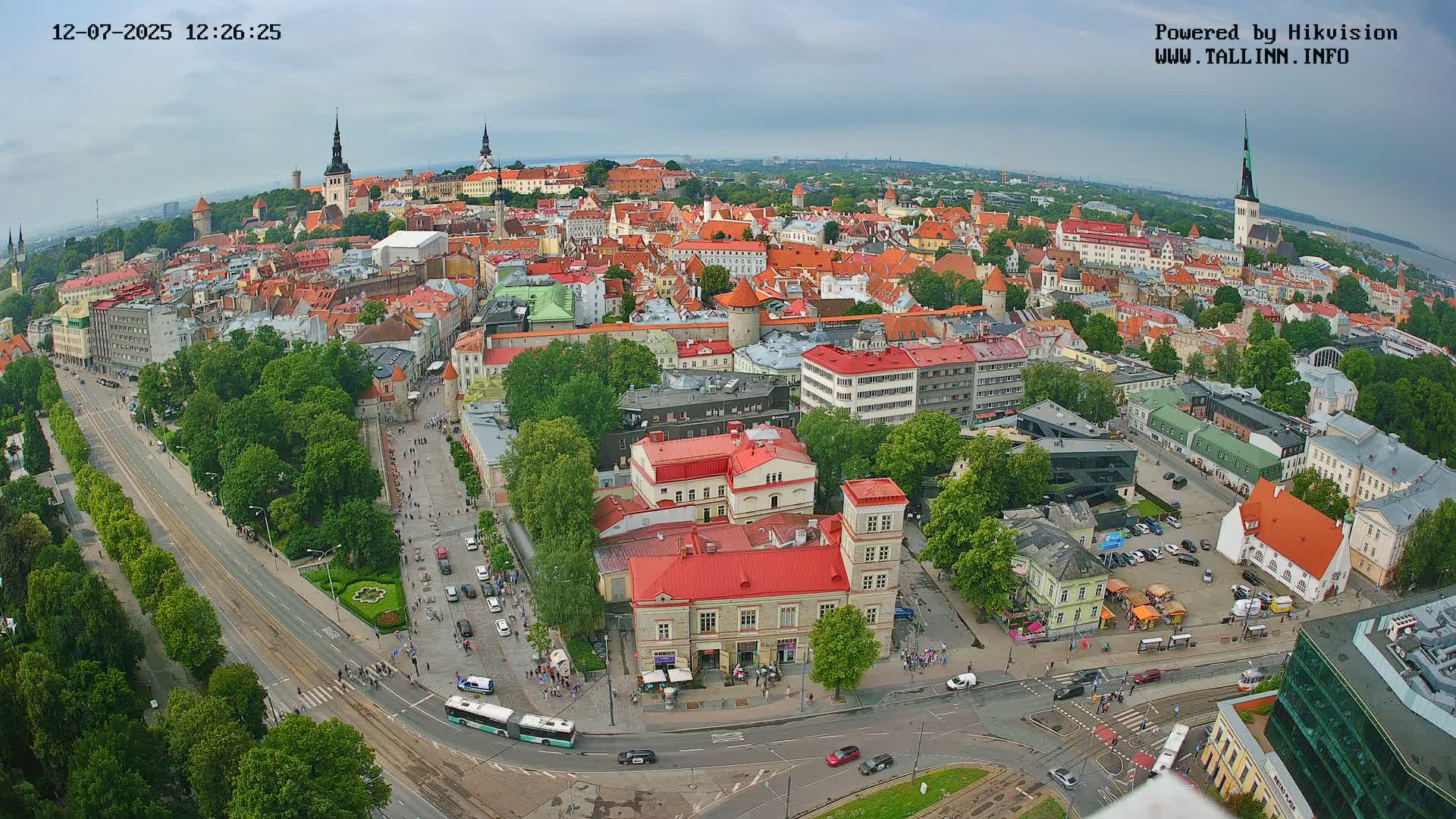 An aerial view of a city on an overcast day shows a large expanse of low buildings with red roofs, several streets with traffic, and green trees.