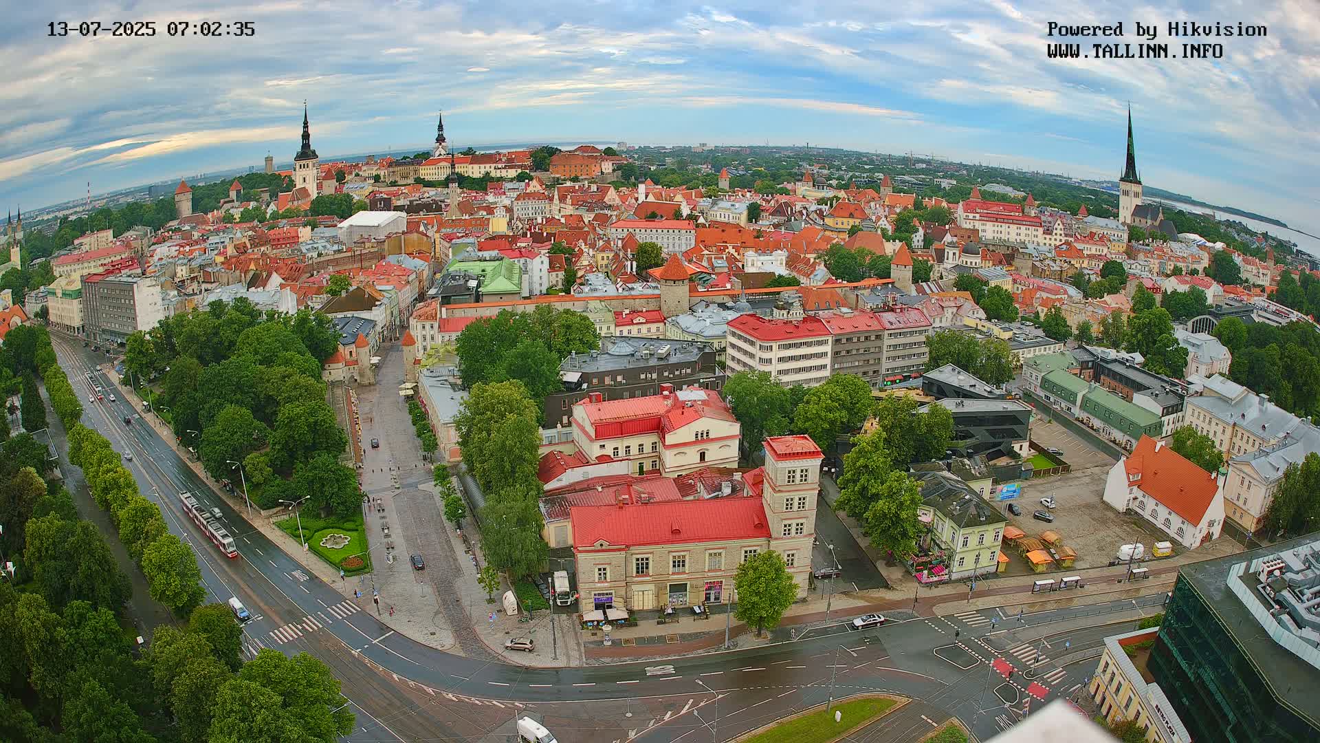 An aerial view of a city on an overcast day shows a large expanse of low buildings with red roofs, several streets with traffic, and green trees.