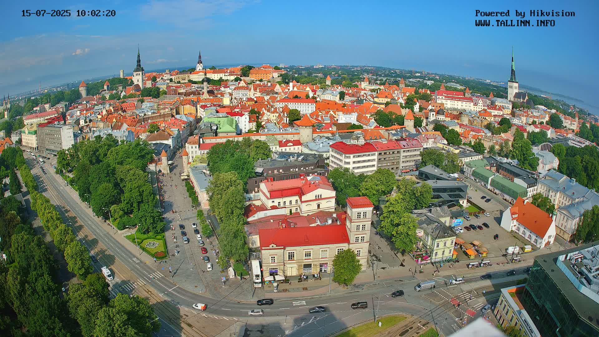 A high-angle, wide view of a city on a sunny day shows numerous red-roofed buildings, tree-lined streets, and a large square.