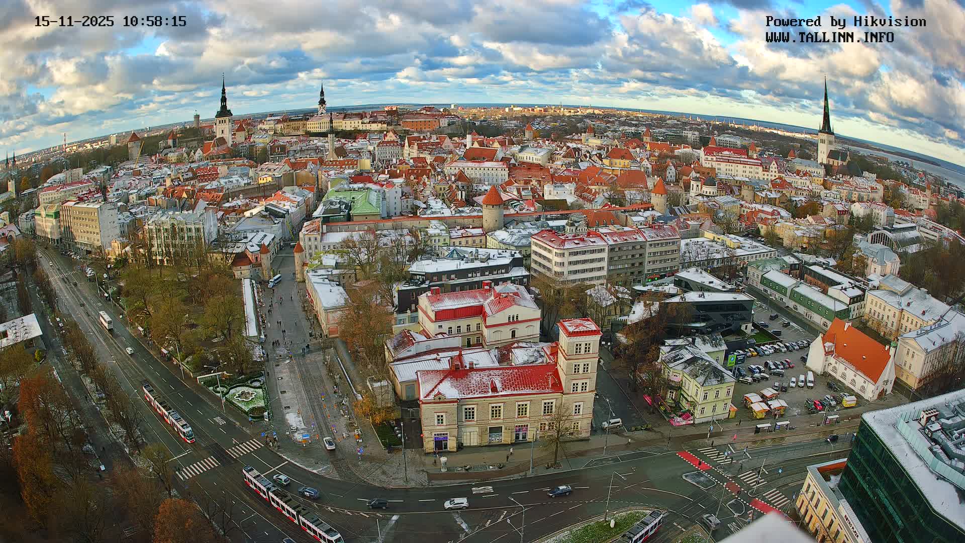 An expansive aerial view reveals a European city featuring a mix of historic red-tiled buildings and church spires dusted with snow, alongside modern streets with trams and cars, all set under a partly cloudy sky with hints of blue.