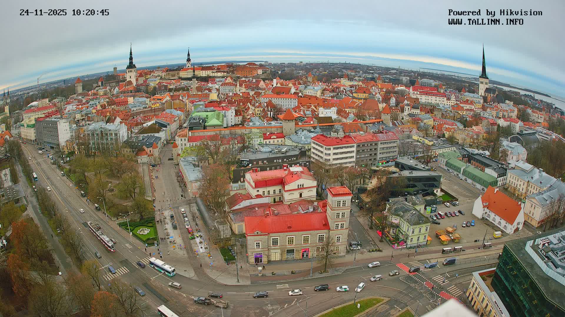 A panoramic high-angle view reveals a bustling historic European city filled with red-roofed buildings, prominent spires, and active streets, all under an overcast sky, with a distant body of water visible on the horizon.