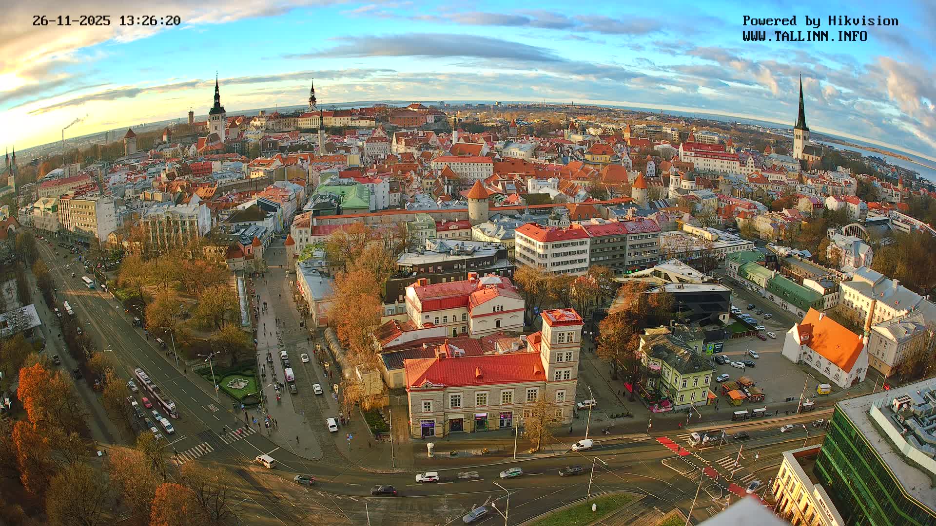 A panoramic high-angle view reveals a bustling historic European city filled with red-roofed buildings, prominent spires, and active streets, all under an overcast sky, with a distant body of water visible on the horizon.