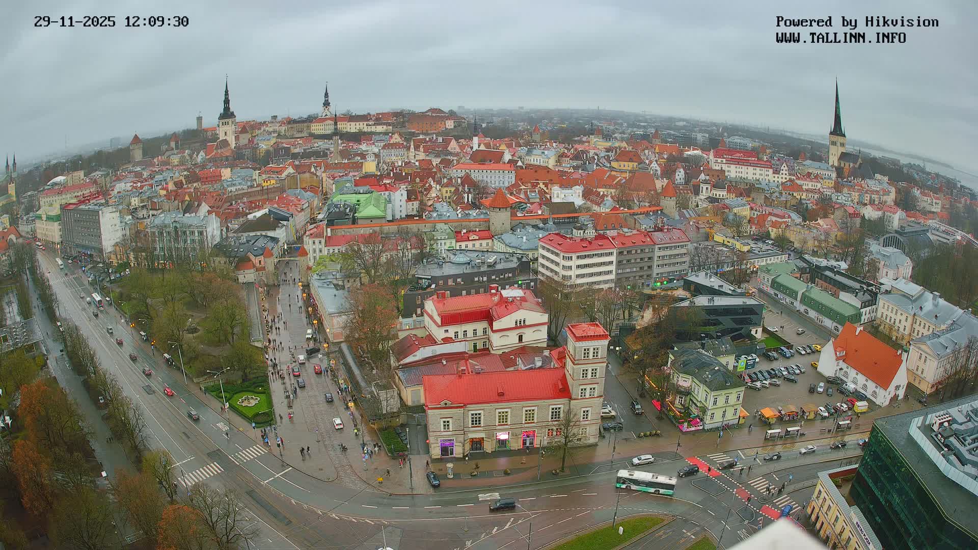 A wide aerial view captures a sprawling, historical European city featuring a dense array of red-roofed buildings, ancient spires, and modern structures, all under a uniformly dull and overcast sky with damp roads, suggesting recent rain.