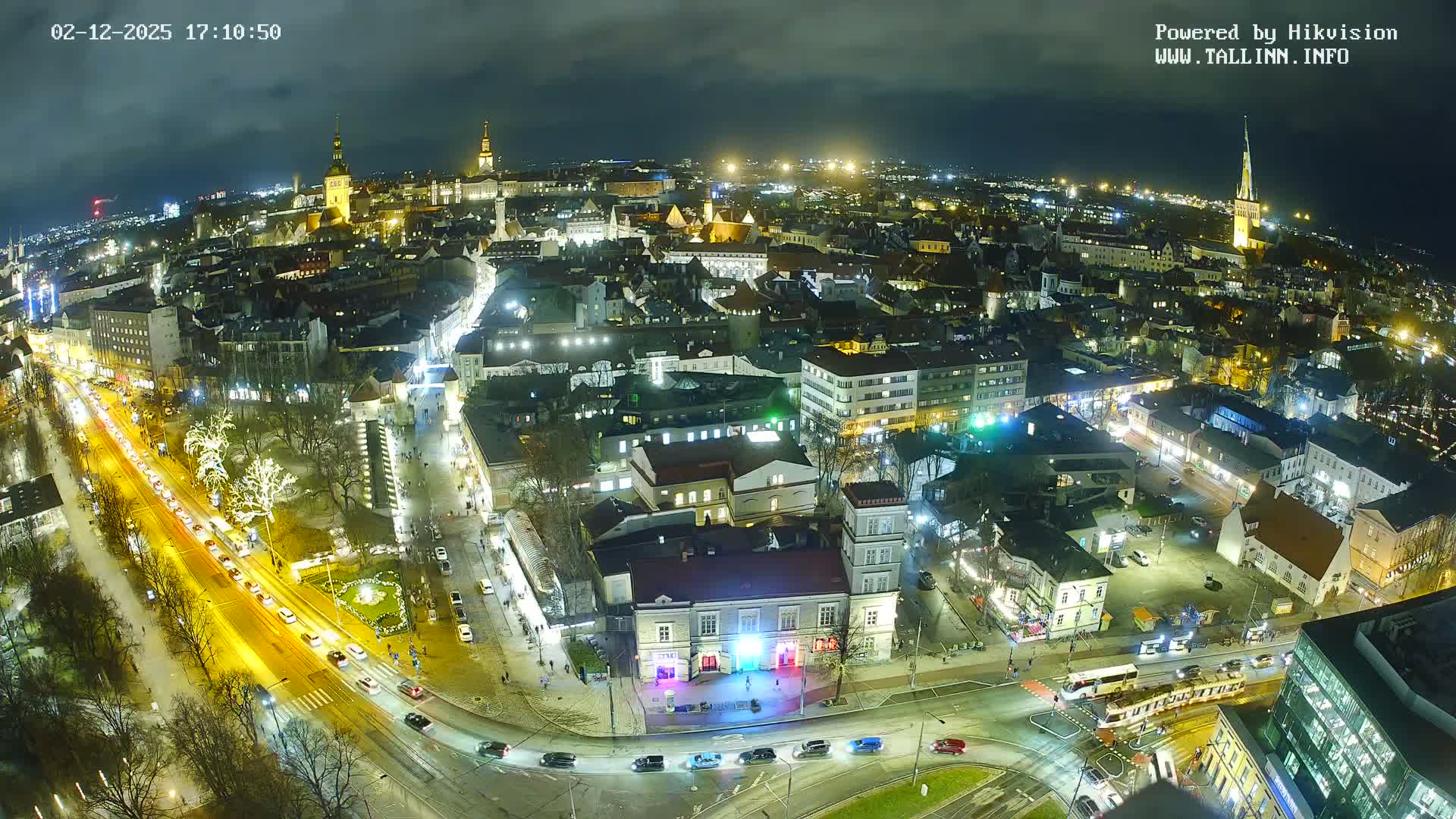 An aerial fisheye view captures a bustling city at night under an overcast sky, illuminated by countless streetlights, building lights, and festive decorations, with cars and a tram moving along busy roads.