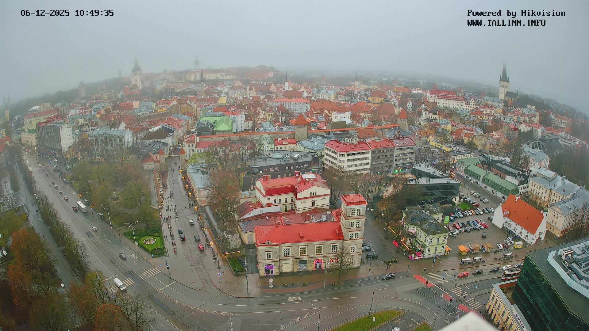 A foggy, overcast day blankets a sprawling European city, revealing a vibrant mix of historical and modern buildings with numerous red roofs, wet streets bustling with traffic and pedestrians, and distant steeples fading into the haze.