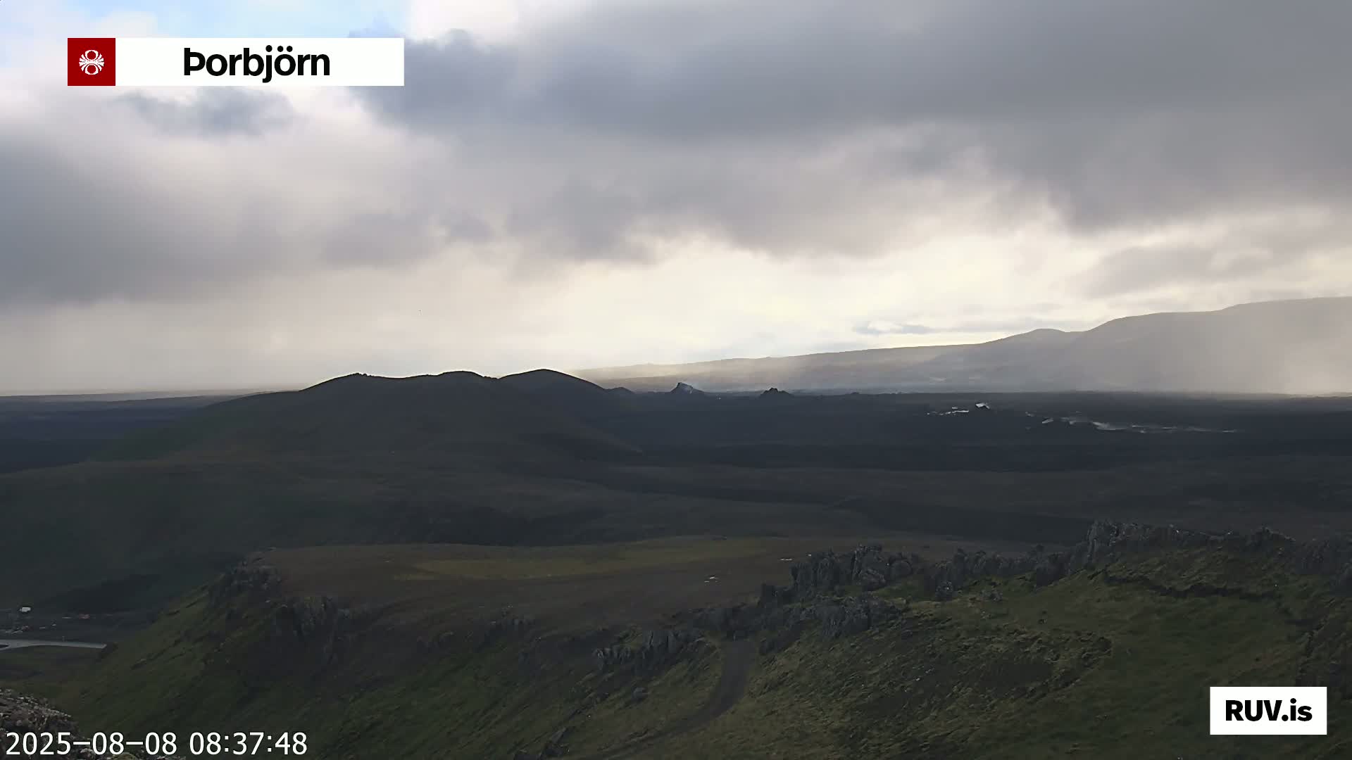 A view of a dark, volcanic landscape under a cloudy, possibly rainy sky shows rolling hills and distant mountains.