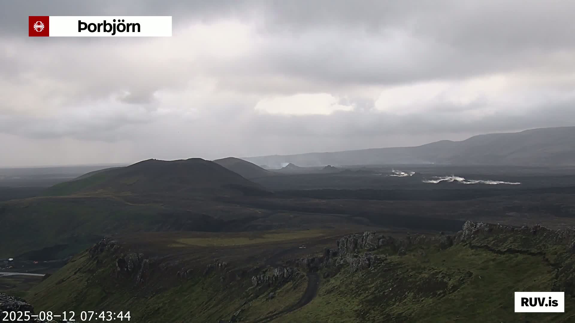 A vast, dark volcanic landscape stretches under a cloudy sky, with hints of steam or smoke rising from a distant area.