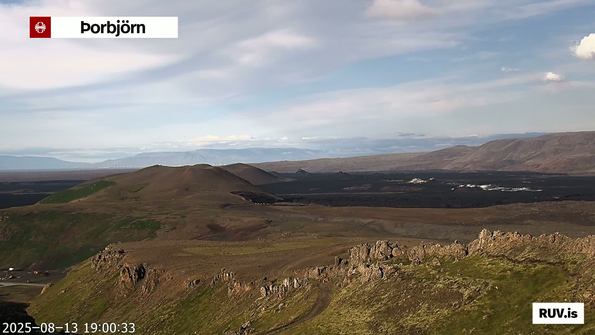 A panoramic view of a volcanic landscape under a partly cloudy sky shows brown hills, dark lava fields, and distant mountains.