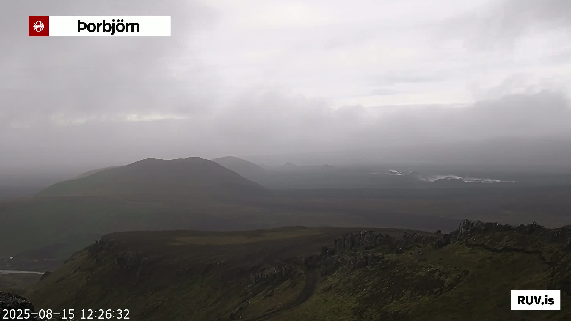 A panoramic view of a volcanic landscape under a partly cloudy sky shows brown hills, dark lava fields, and distant mountains.