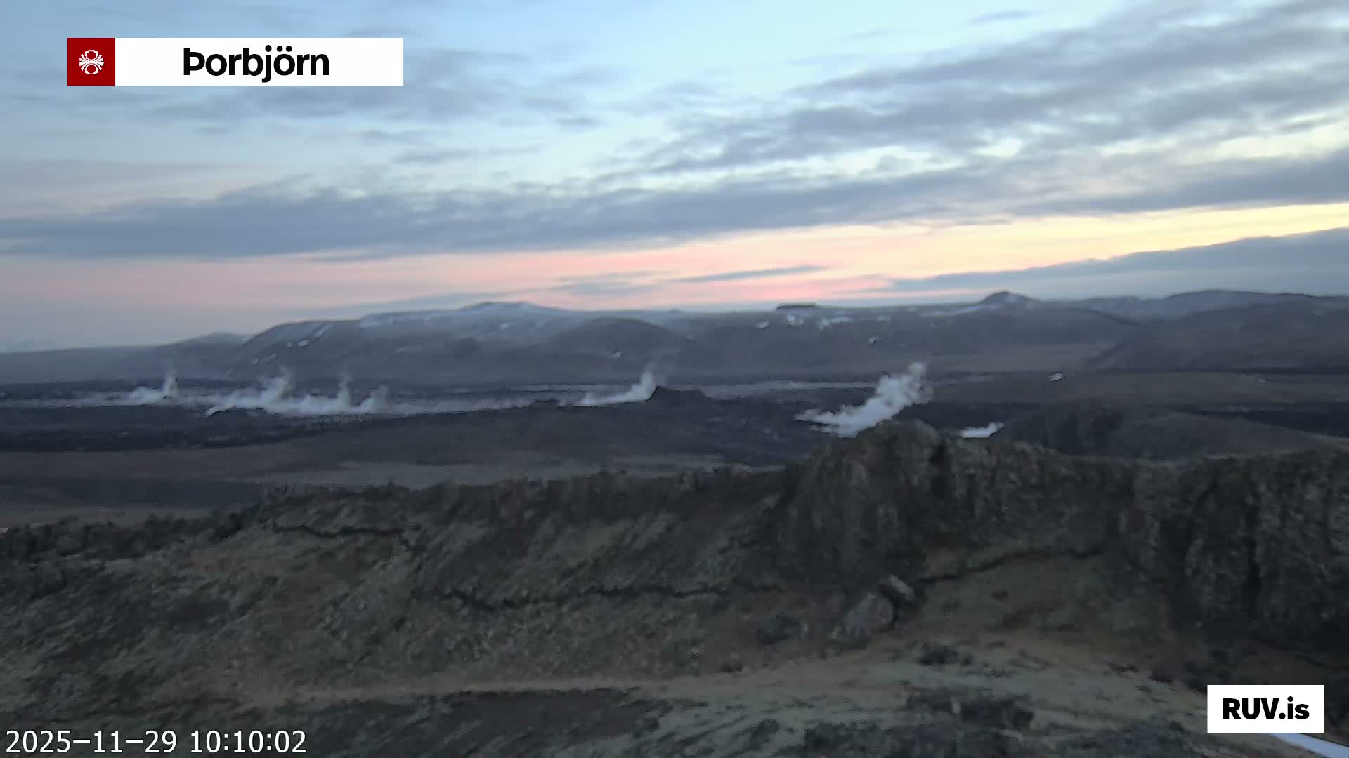 Under an overcast sky with soft pink and orange hues, a rugged, dark volcanic landscape stretches out, featuring several distinct plumes of white steam rising from the ground towards distant snow-dusted mountains.