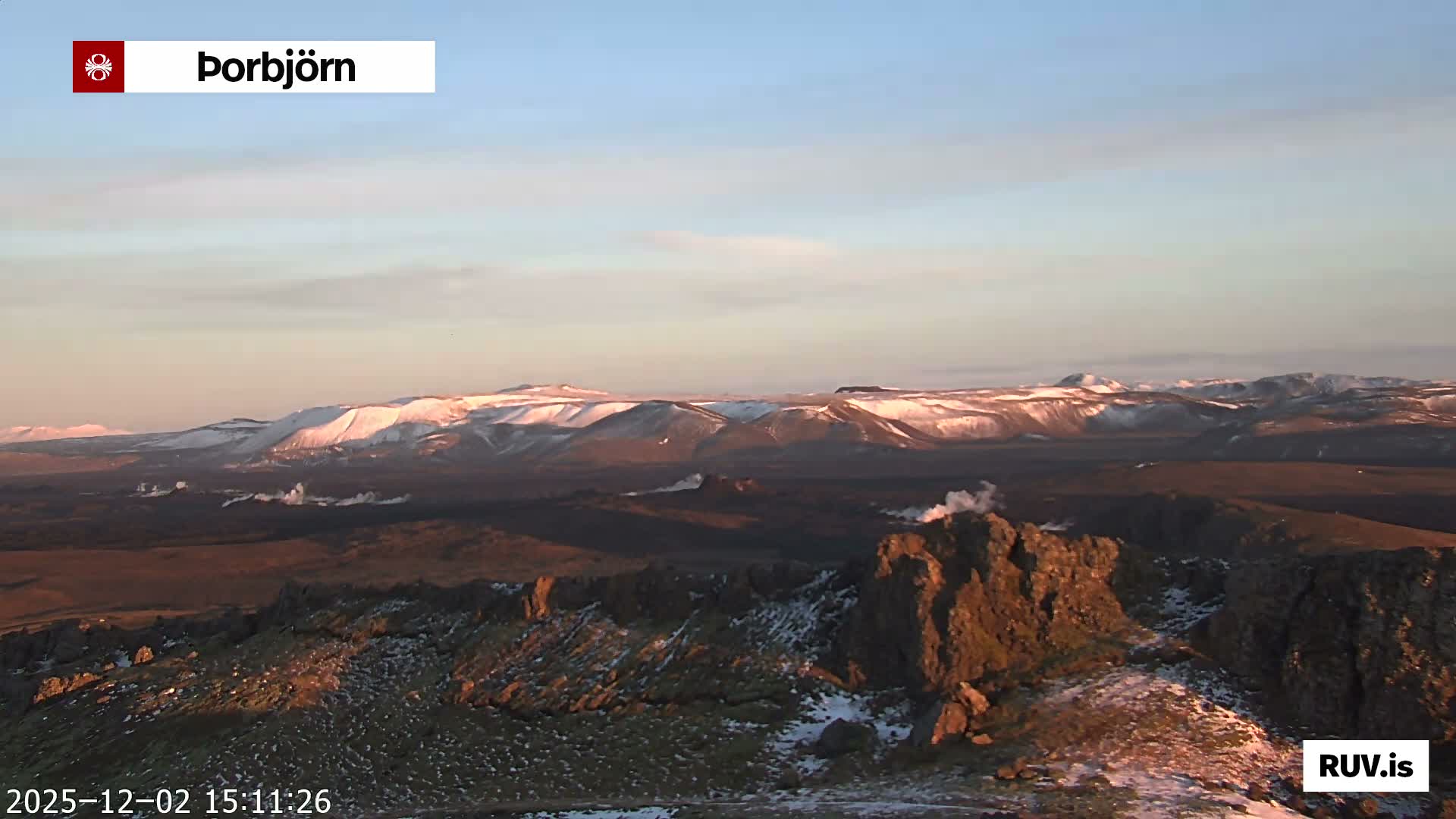 A cold, rugged landscape with patches of snow and dark ground features several plumes of steam rising, stretching towards distant snow-covered mountains under a clear sky illuminated by low, warm sunlight.