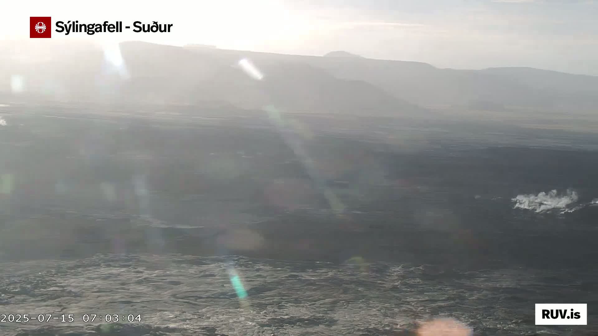 A hazy, sunlit view of a dark ocean with distant mountains under a mostly clear sky shows small plumes of water in the distance.