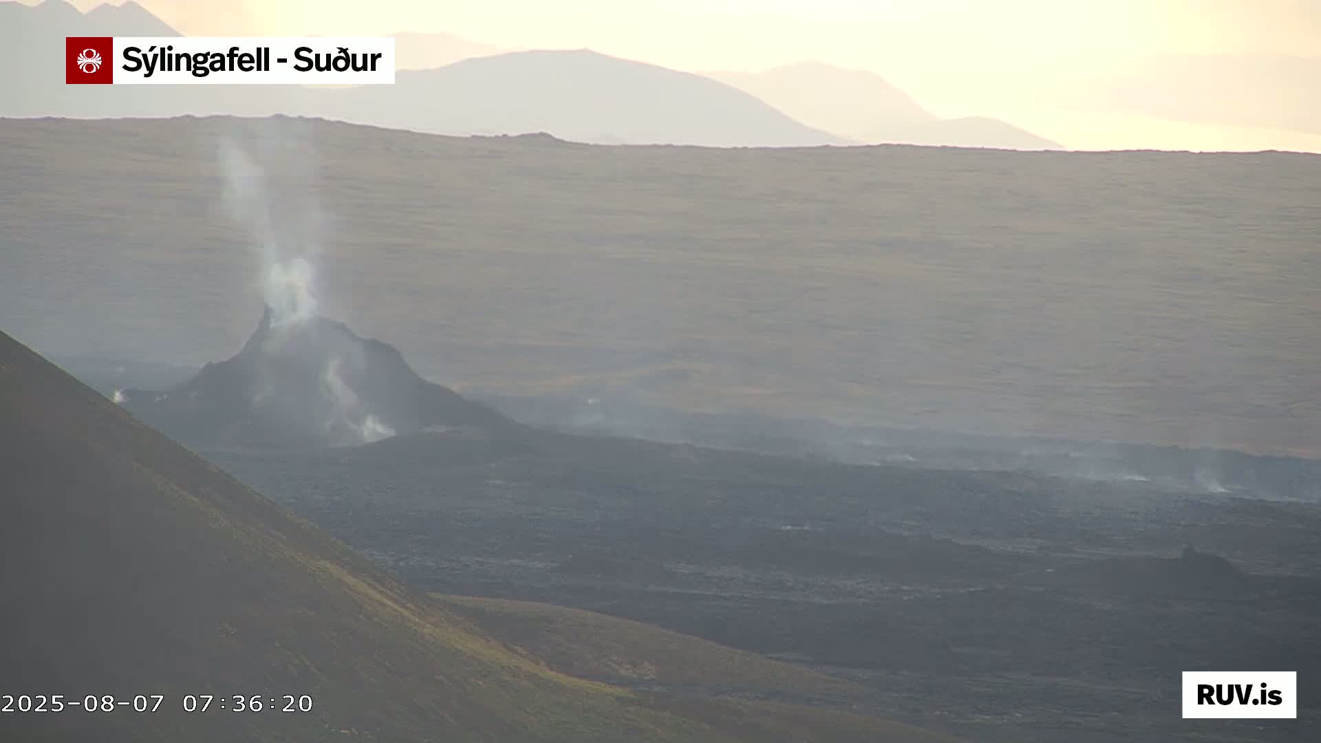 A volcanic eruption is seen from a distance, with a plume of smoke rising from a dark, cone-shaped vent, under hazy, light conditions.