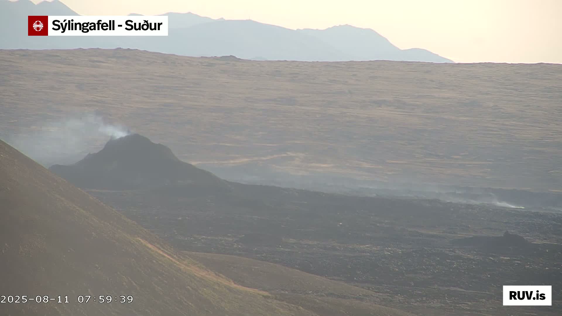 A volcanic landscape under hazy conditions shows a dark, smoking cone in the foreground, with a vast expanse of dark brown and gray terrain extending to distant, faintly visible mountains.