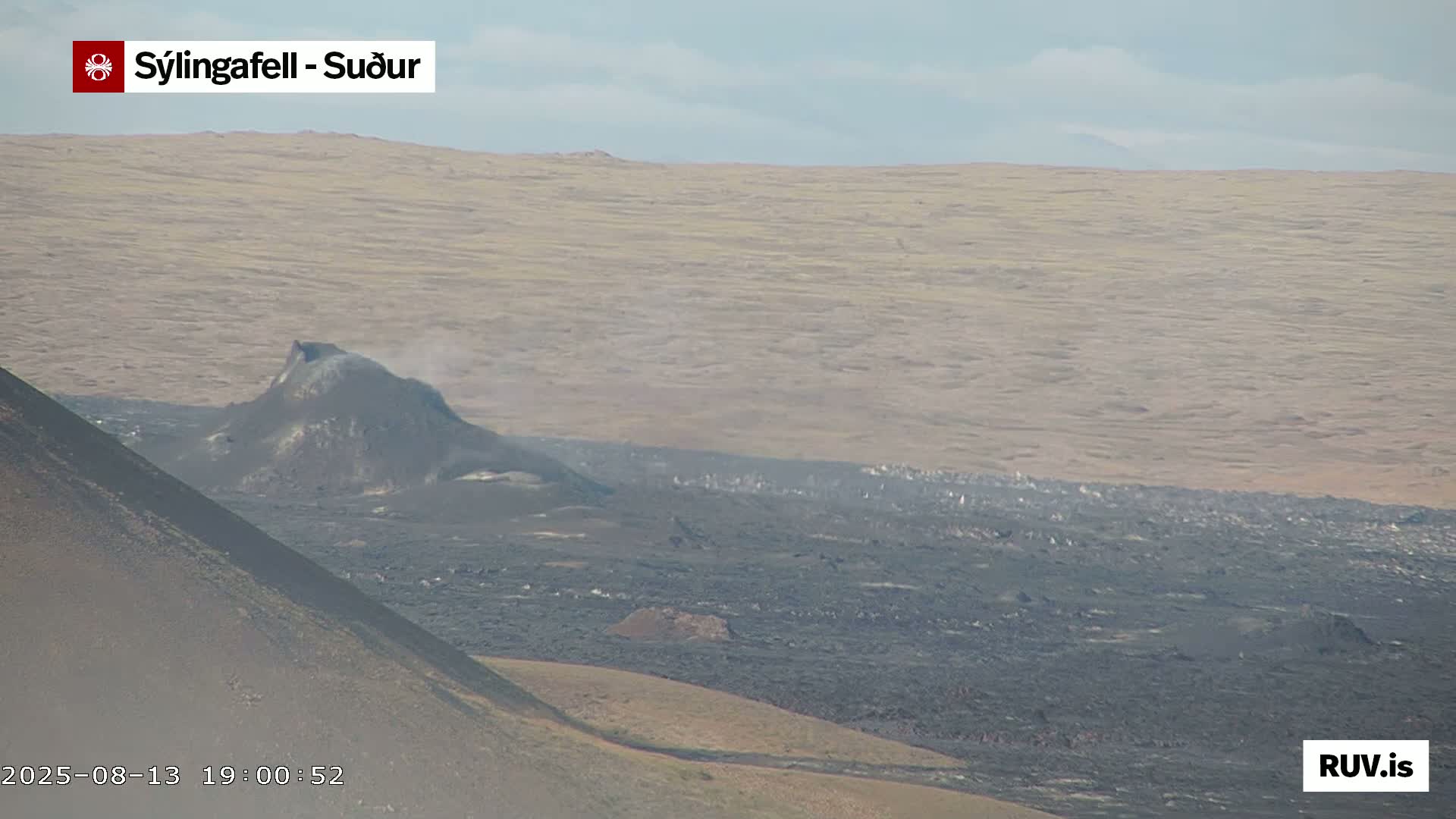 A dark volcanic cone with a small plume of smoke is seen amidst a vast expanse of dark, solidified lava, under a hazy sky.