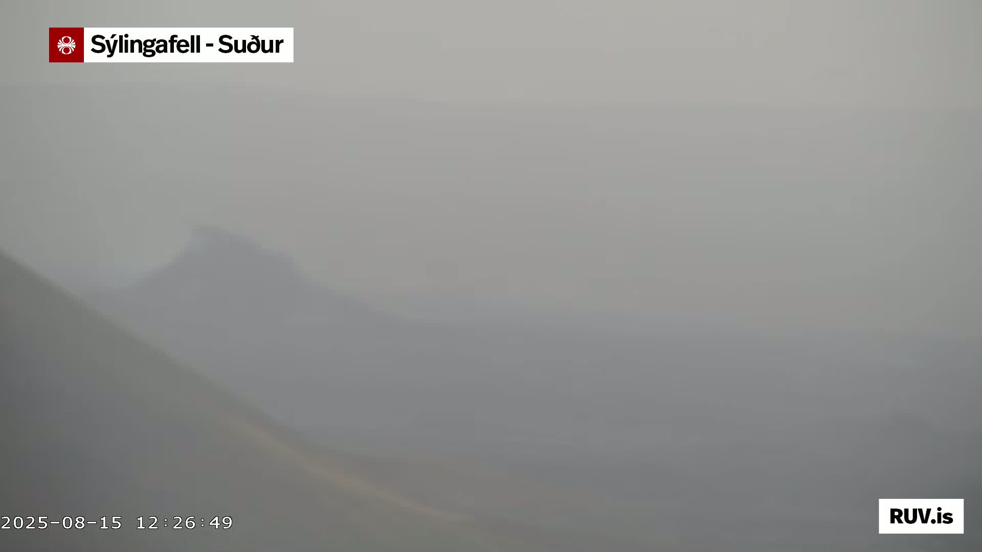 A dark volcanic cone with a small plume of smoke is seen amidst a vast expanse of dark, solidified lava, under a hazy sky.