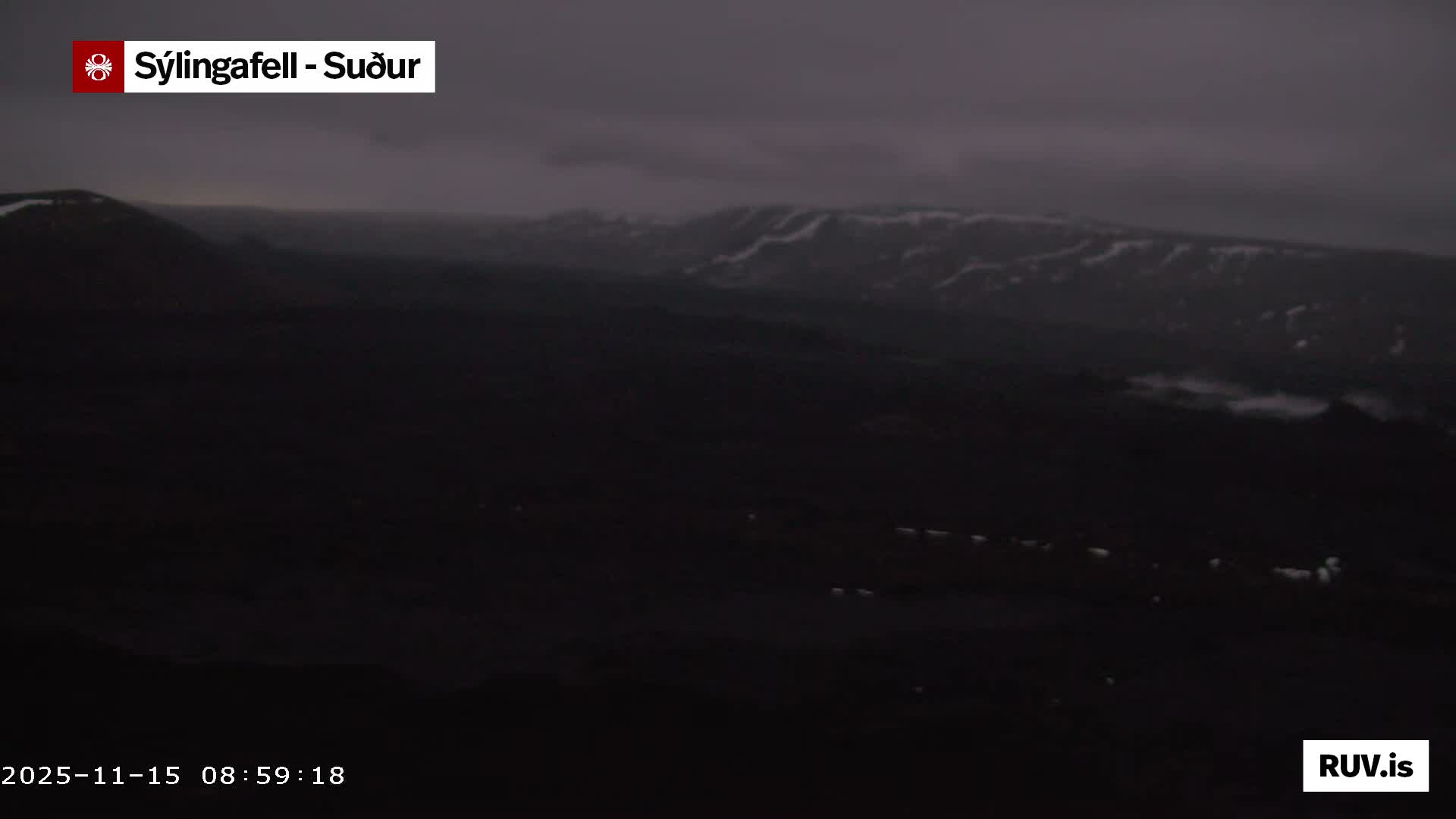 The image displays a dark, expansive mountainous landscape under a heavily overcast and gloomy sky, with faint patches of snow or light visible on distant peaks and some low-lying fog in the valleys.
