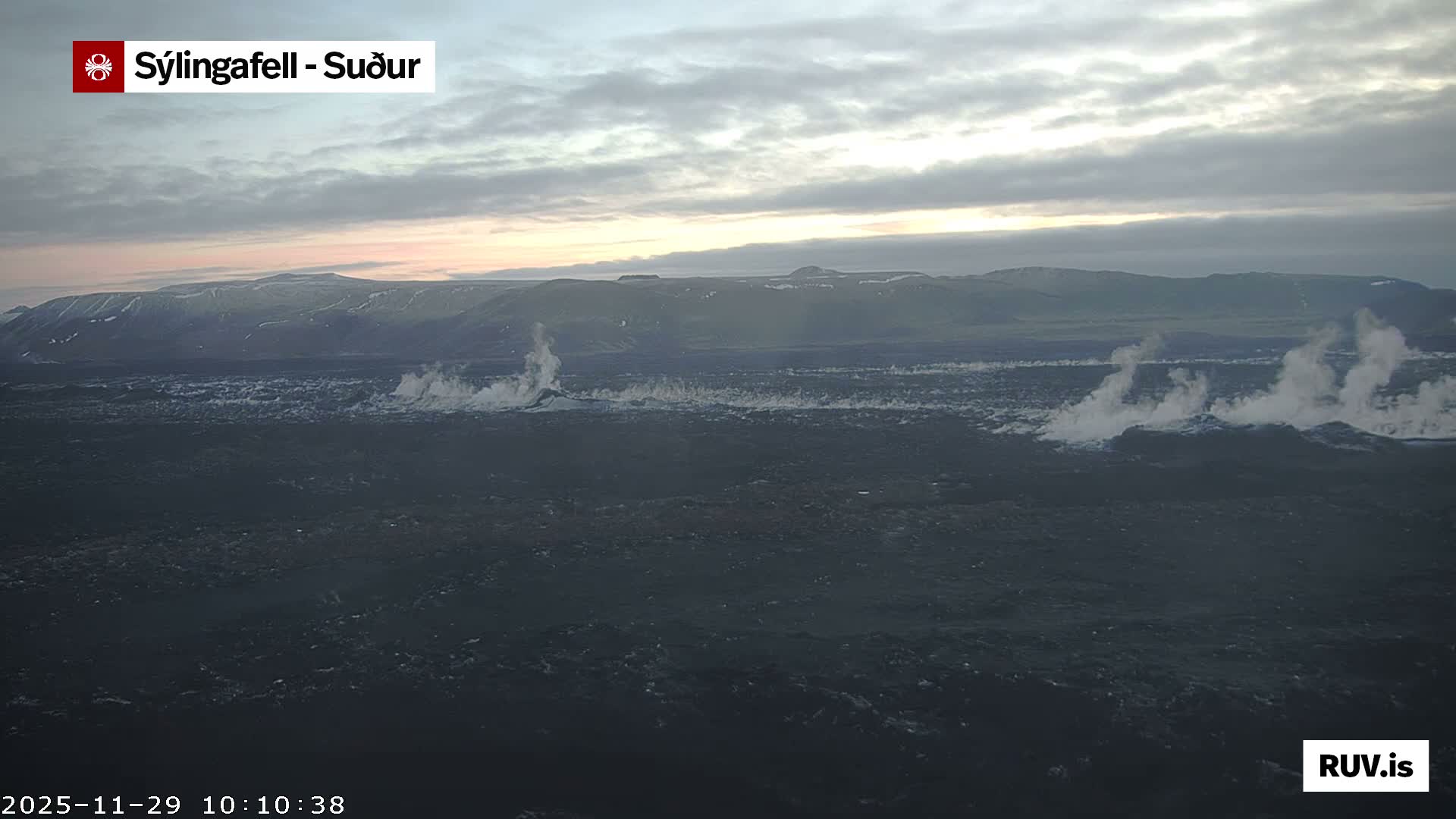 A rugged, dark volcanic landscape features multiple plumes of white steam rising from the ground, with distant mountains under a cloudy sky displaying subtle dawn or dusk colors.