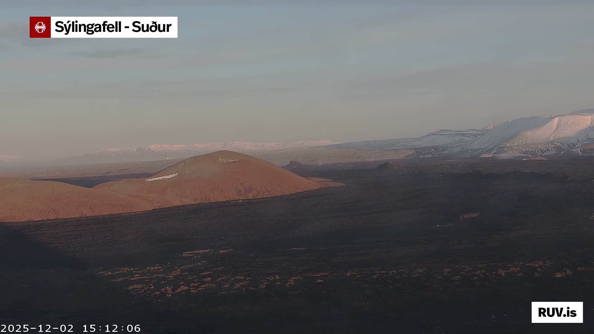 A cold, hazy day over a volcanic landscape features dark lava fields, a reddish-brown cinder cone dusted with snow, and snow-capped mountains in the distance.