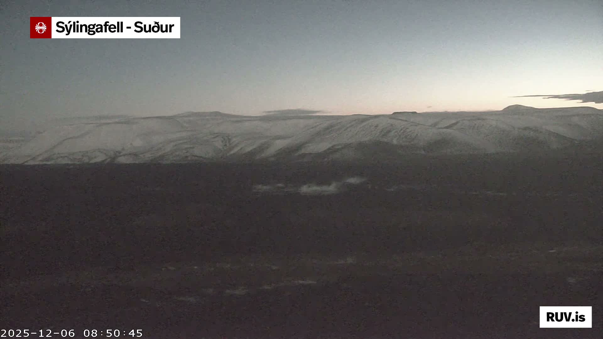 A vast, dark, snow-covered mountain landscape is depicted under a dim, largely clear sky with hints of dawn or dusk on the horizon, featuring patches of low-lying fog in the dark foreground.