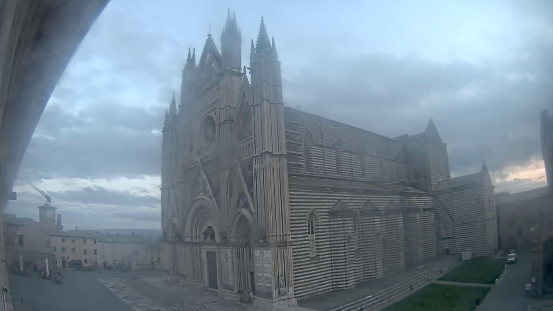 A large, striped cathedral stands in a town square on an overcast day, with a light mist or fog obscuring parts of the scene.