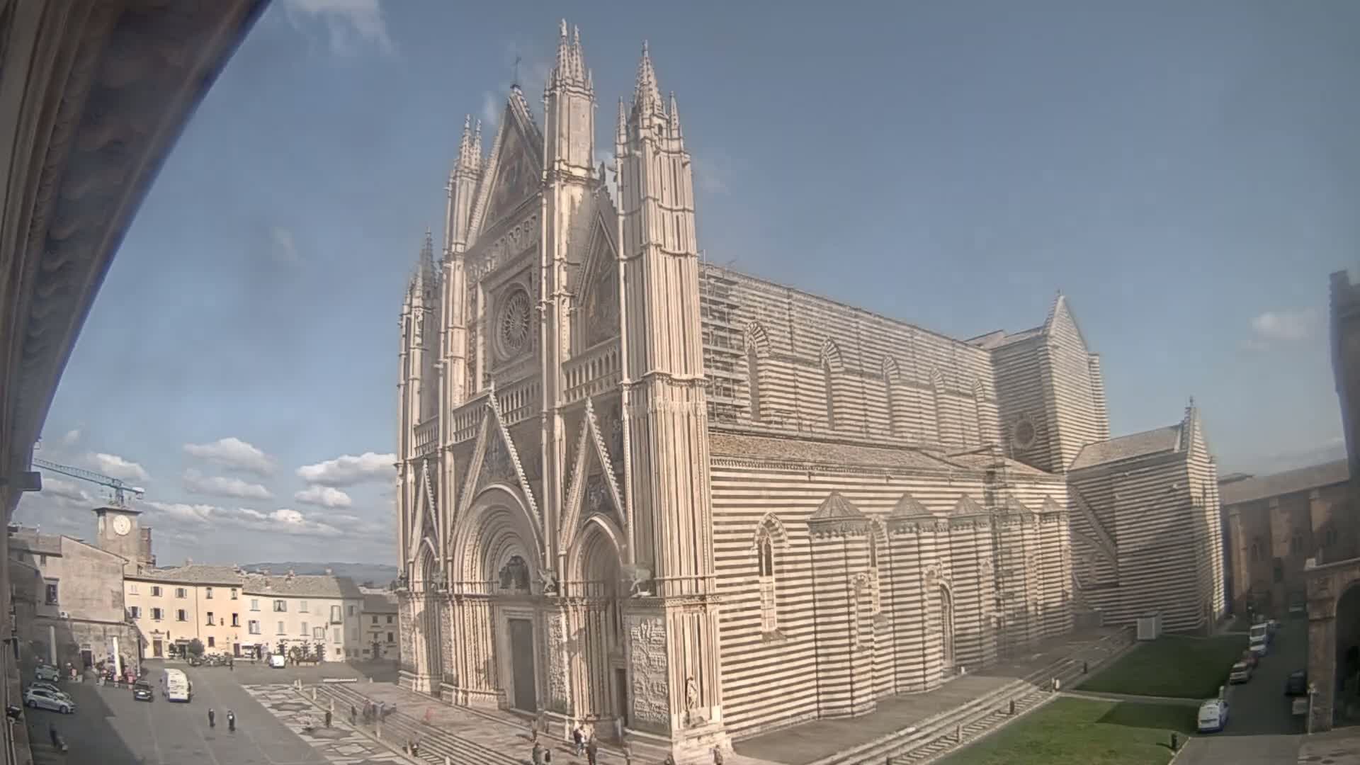 A large, striped stone cathedral stands in a city square under a partly cloudy sky.