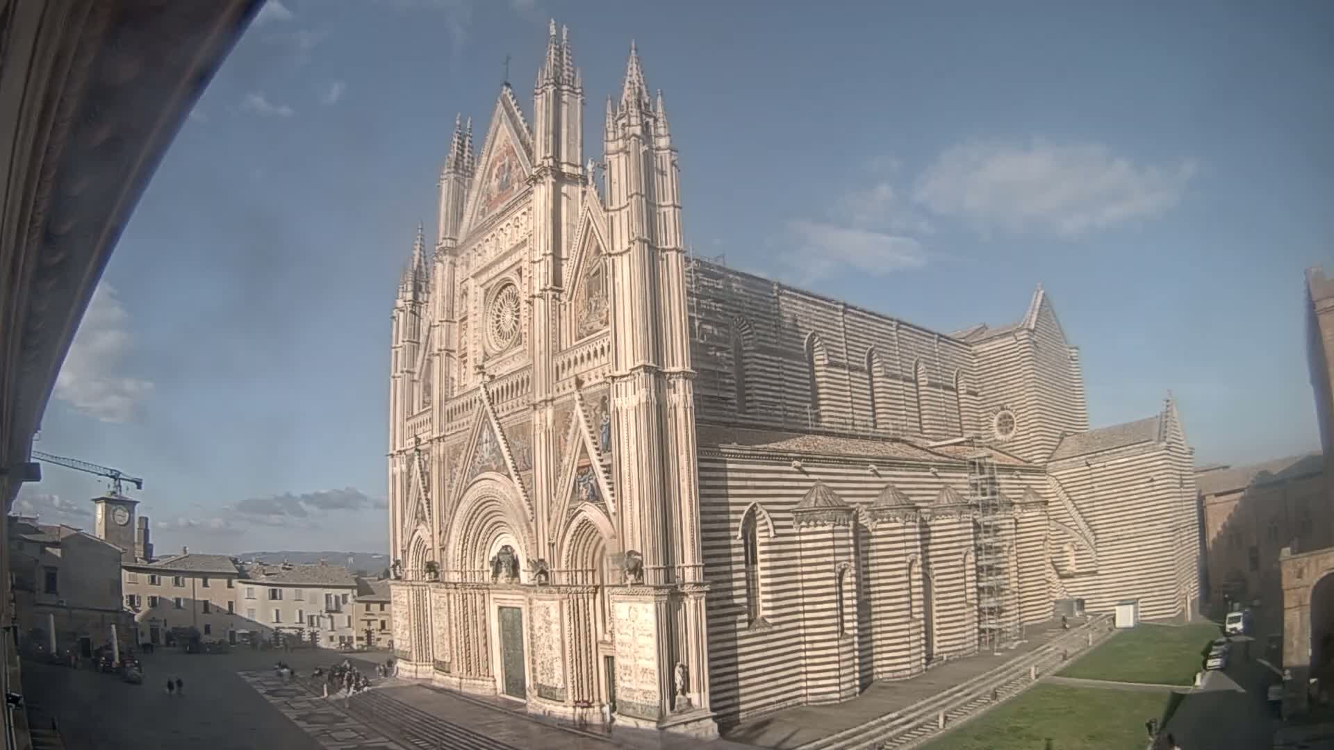 A large, striped cathedral with Gothic architecture stands in a city square under a mostly sunny sky.
