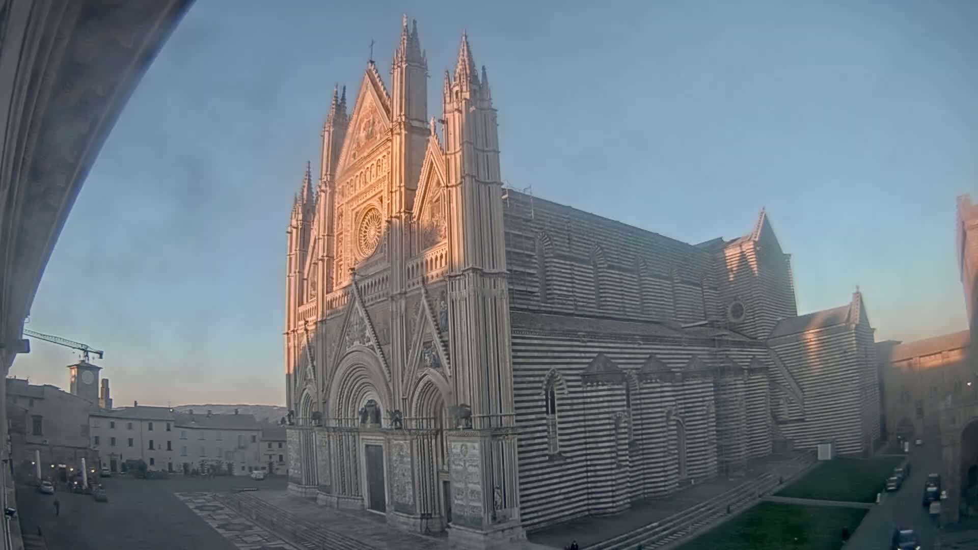 A large, striped cathedral stands in a town square on a clear, sunrise-lit morning.