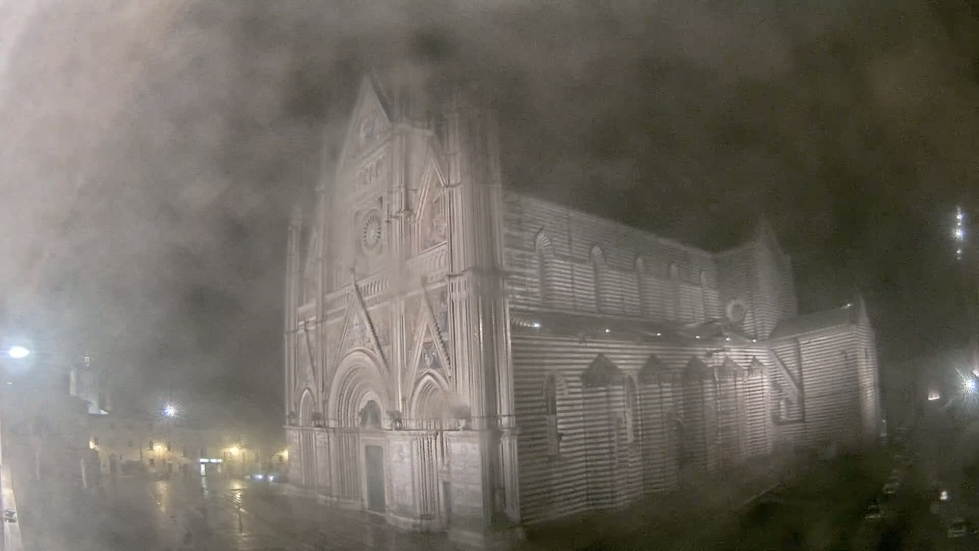 A large, striped stone cathedral stands at night in heavy fog and rain.