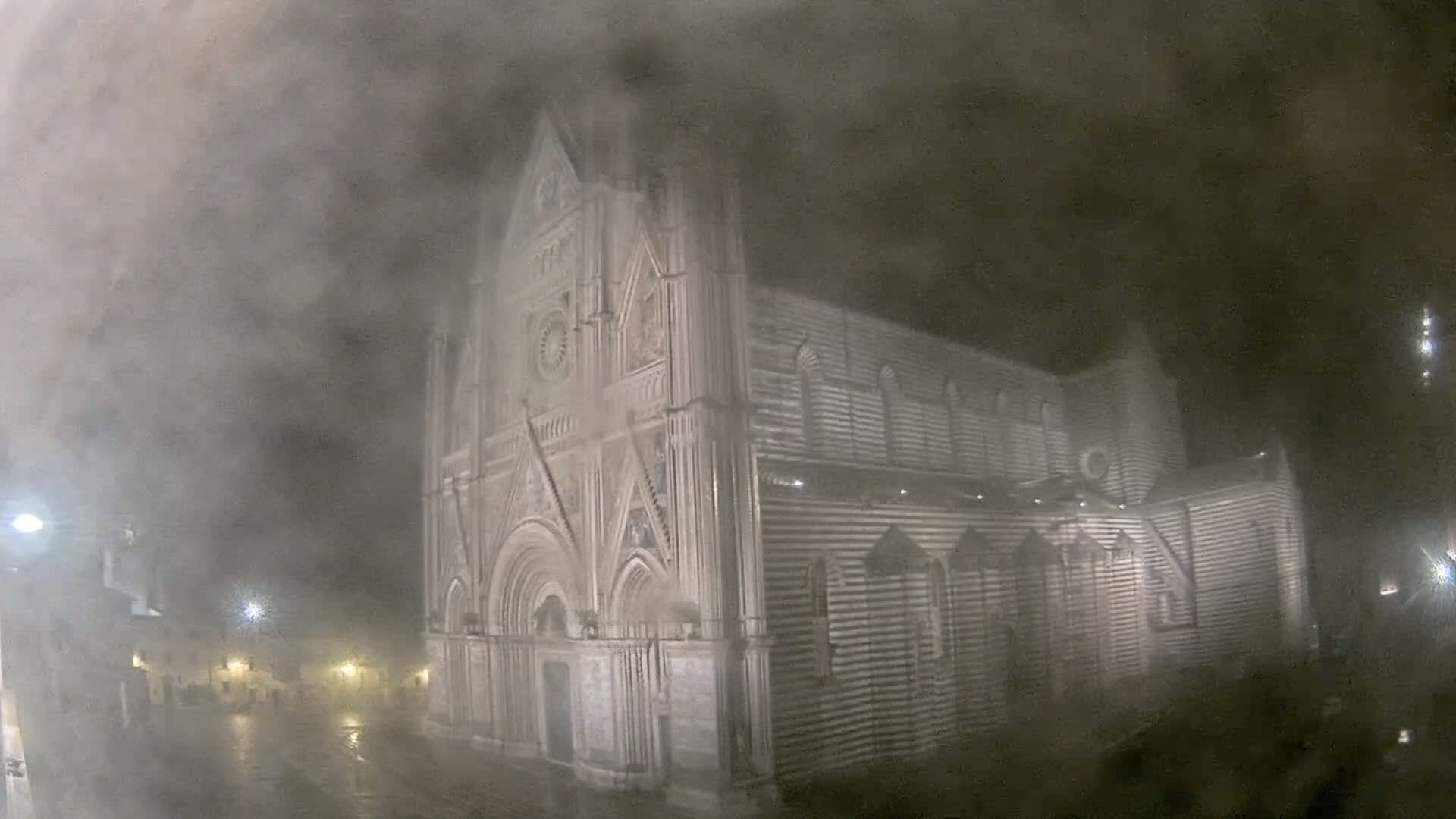 A large, striped stone cathedral is visible at night in heavy fog or mist, with some streetlights illuminating a wet, empty plaza in the foreground.