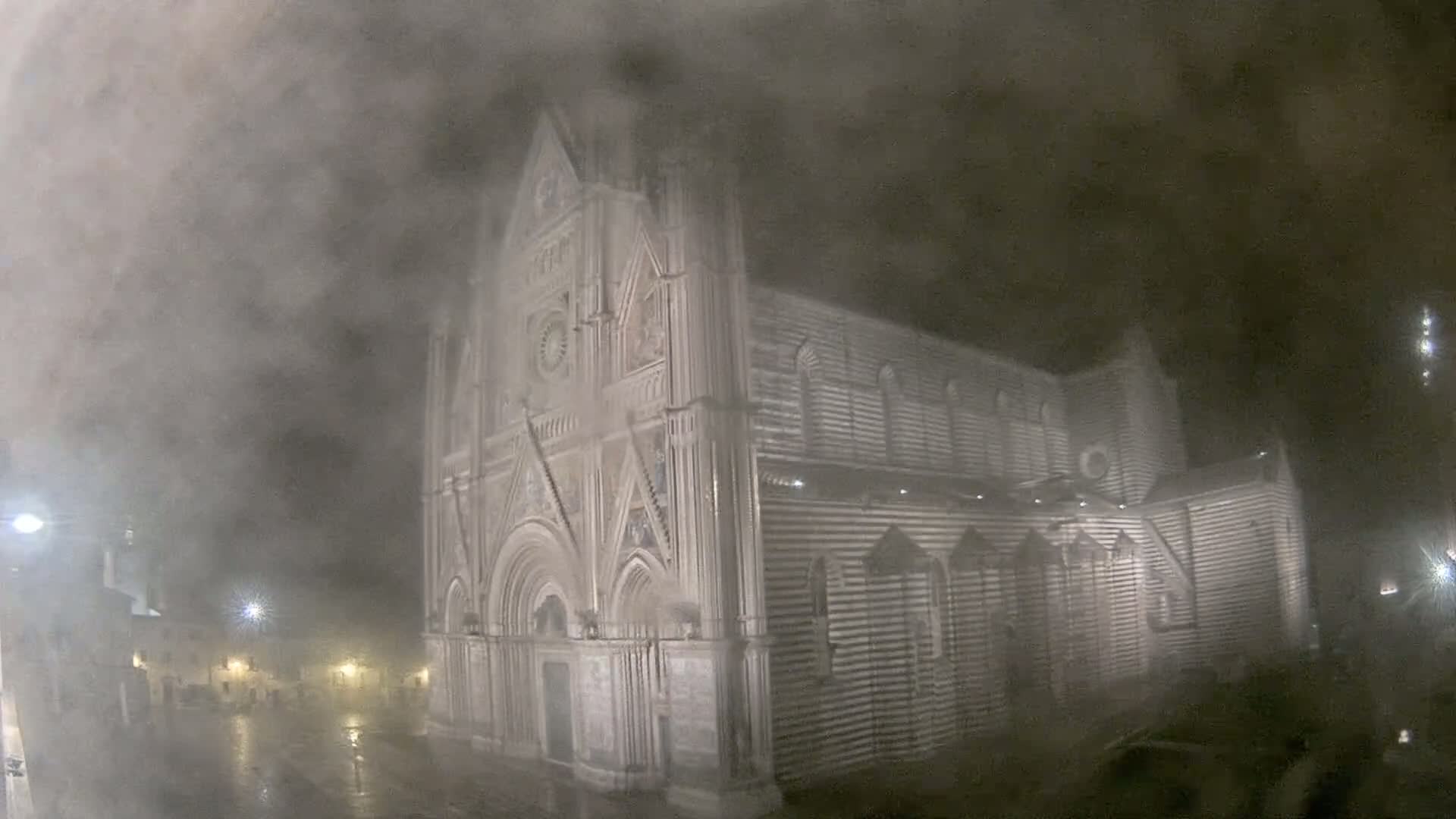 A large, striped stone cathedral stands at night in a heavy fog and rain.