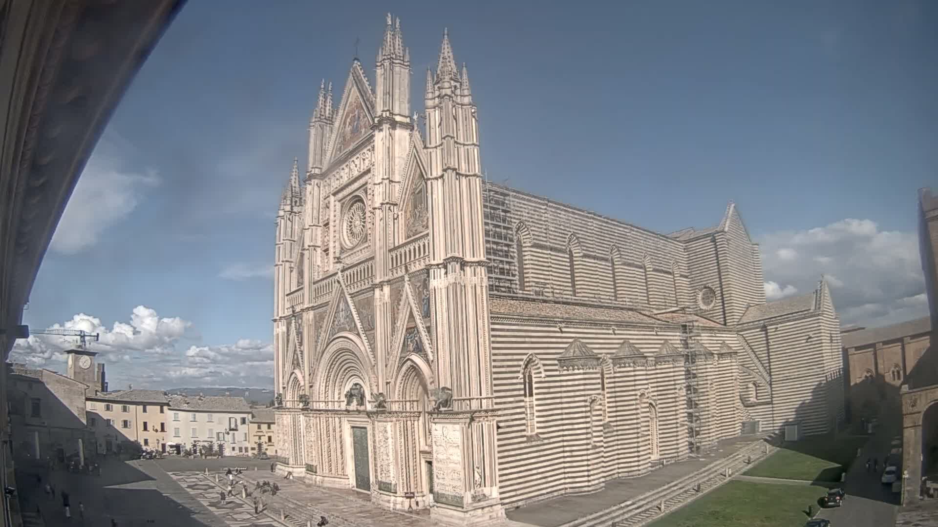 A large, striped cathedral with Gothic-style architecture stands in a town square under a partly cloudy sky.