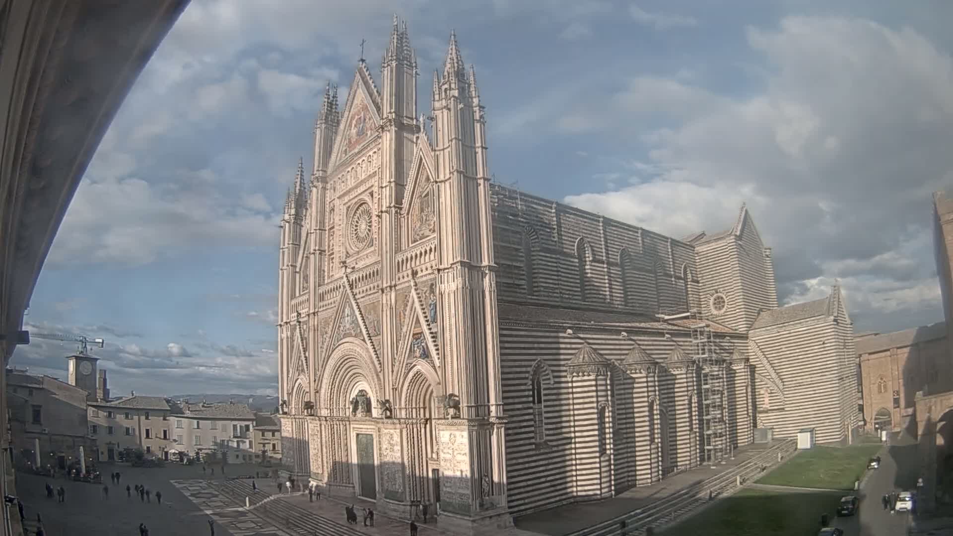 A large, striped cathedral stands in a town square under a partly cloudy sky.