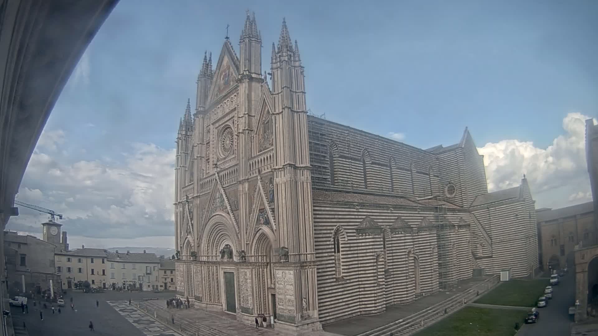 A large, striped stone cathedral stands in a town square under a partly cloudy sky.