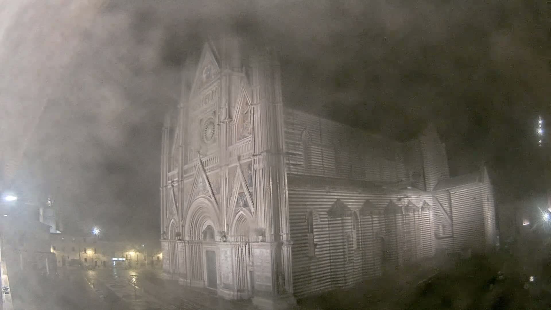 A large, striped stone cathedral stands in a town square at night, obscured by heavy fog or mist in the rain.