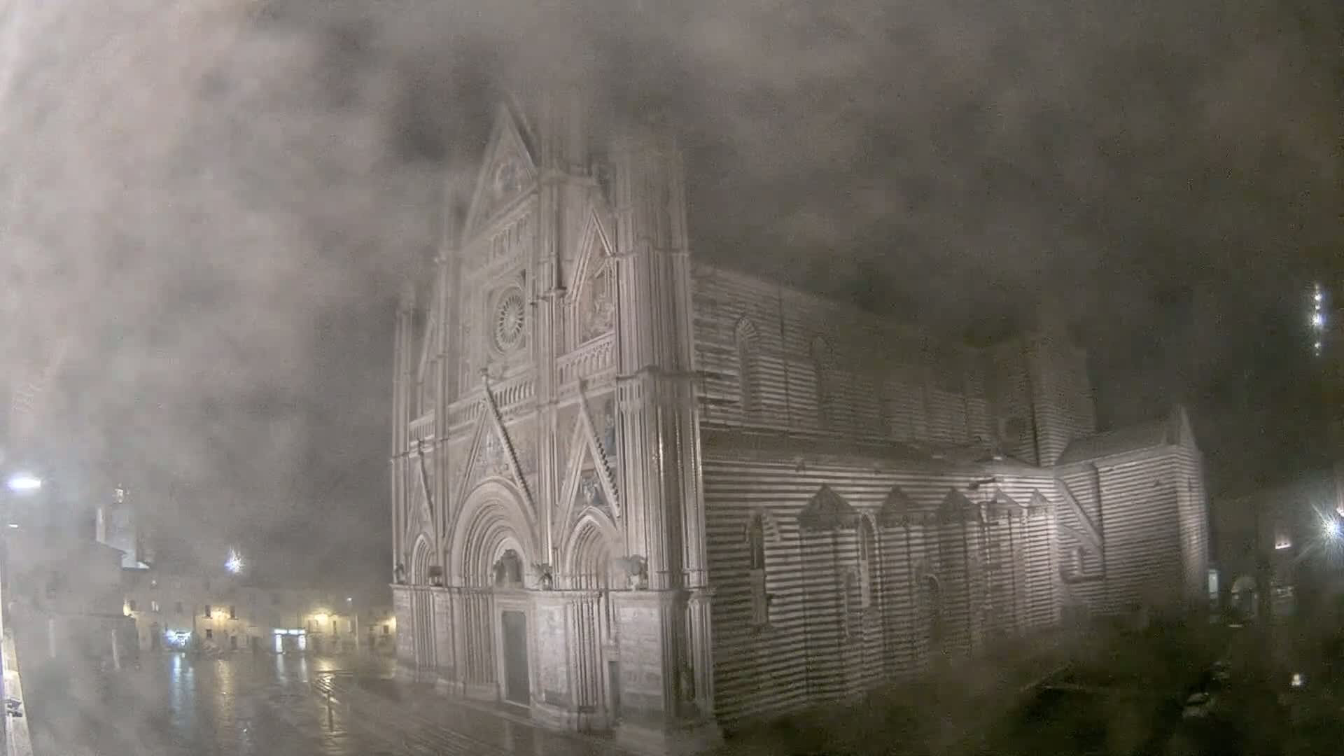 A large, striped cathedral stands in a town square shrouded in heavy fog and rain at night.