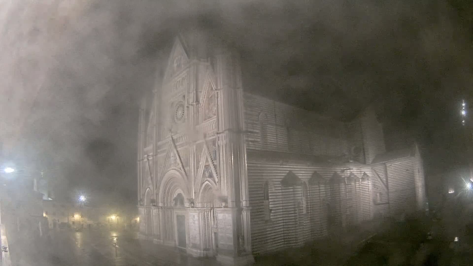 A large, ornate stone building, possibly a cathedral, is shrouded in thick fog or mist under nighttime conditions, with rain visible on the ground.