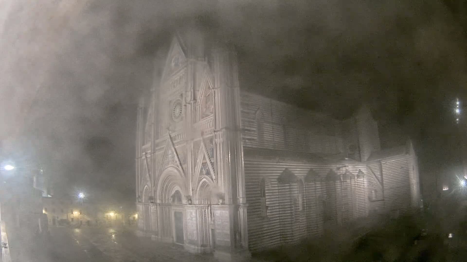 A large stone cathedral is partially obscured by heavy fog or mist at night, with faint streetlights visible in the background.