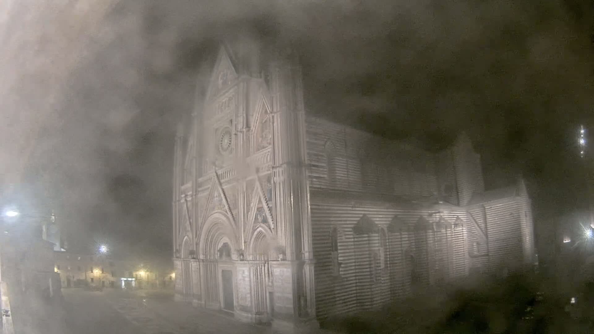 A large, striped stone cathedral stands in a town square at night, shrouded in heavy fog or mist.