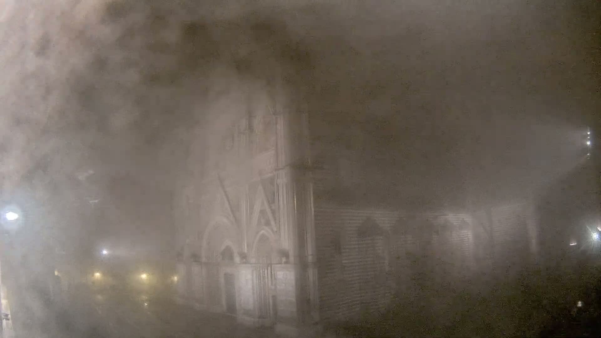 A stone building is partially obscured by a thick fog at night, with faint lights visible in the distance.