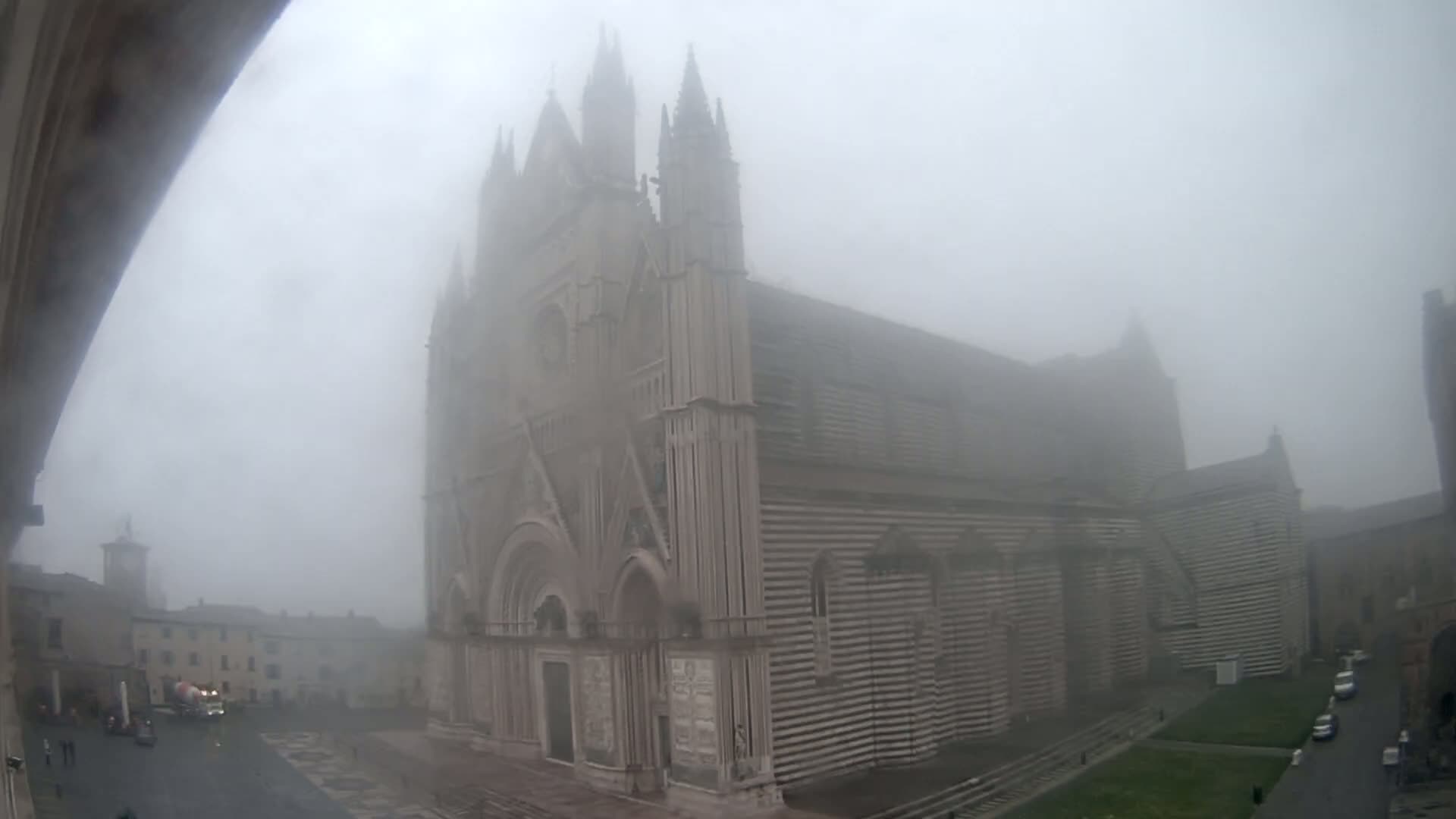 A large stone cathedral, partially obscured by dense fog, stands in a town square with a few vehicles and buildings visible in the background.