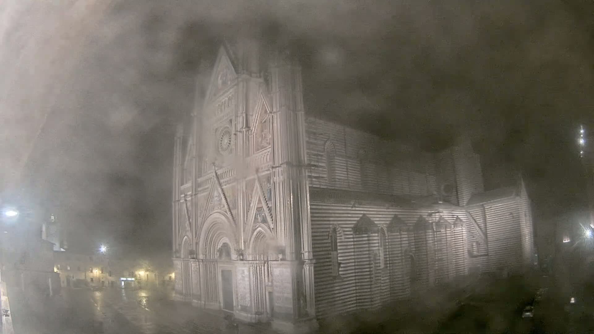 A large, striped stone cathedral stands in a town square at night, obscured by heavy fog and rain.