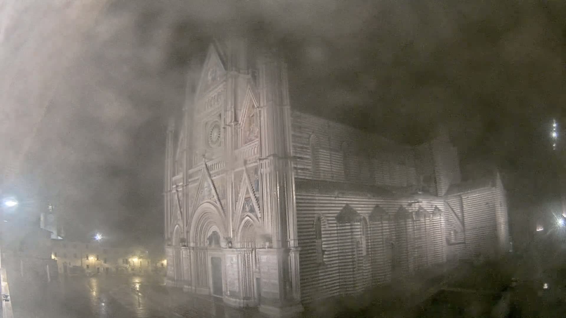 A large, striped stone cathedral stands at night in heavy fog and rain.
