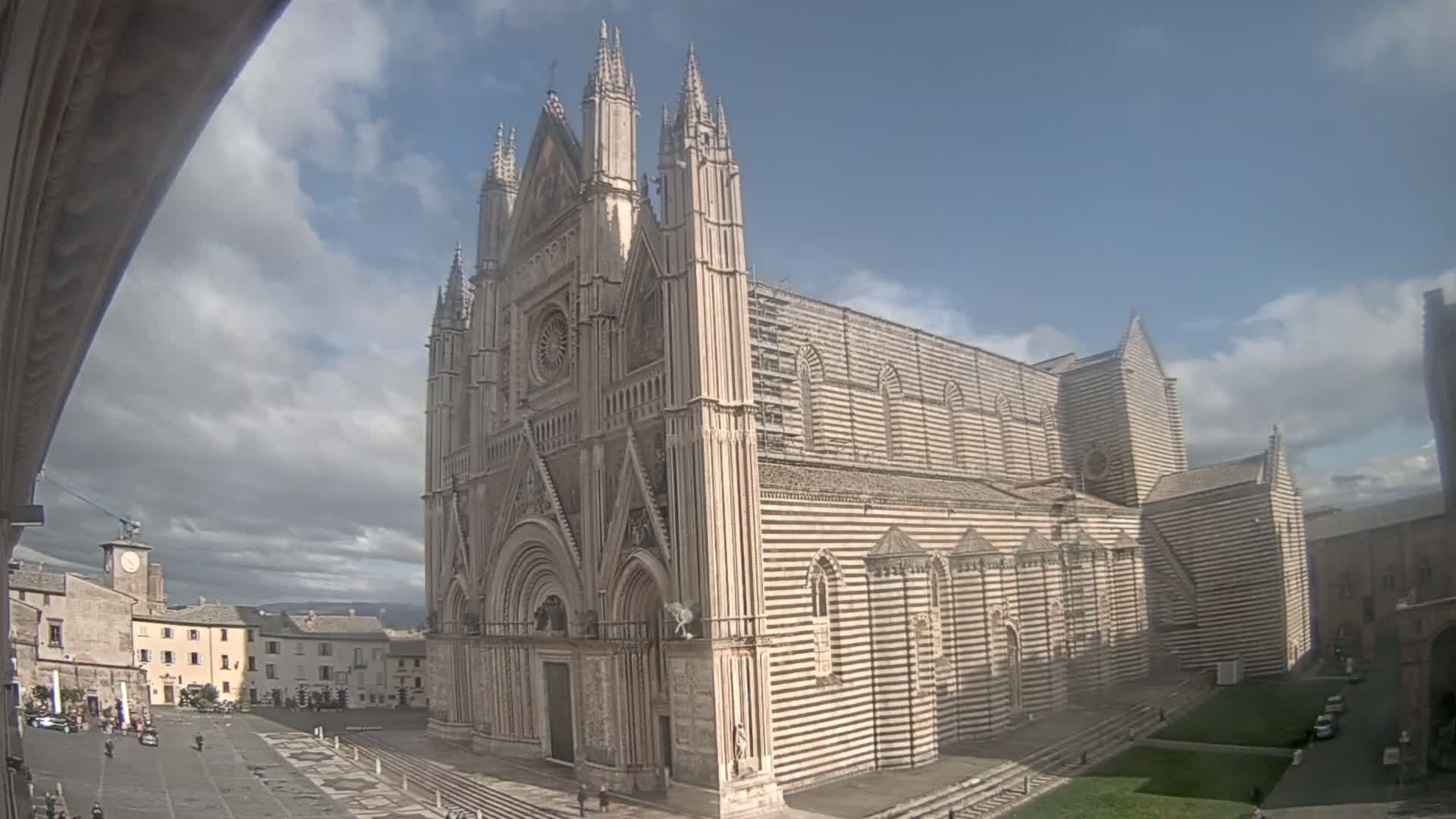 A large, striped stone cathedral stands in a town square under a partly cloudy sky.