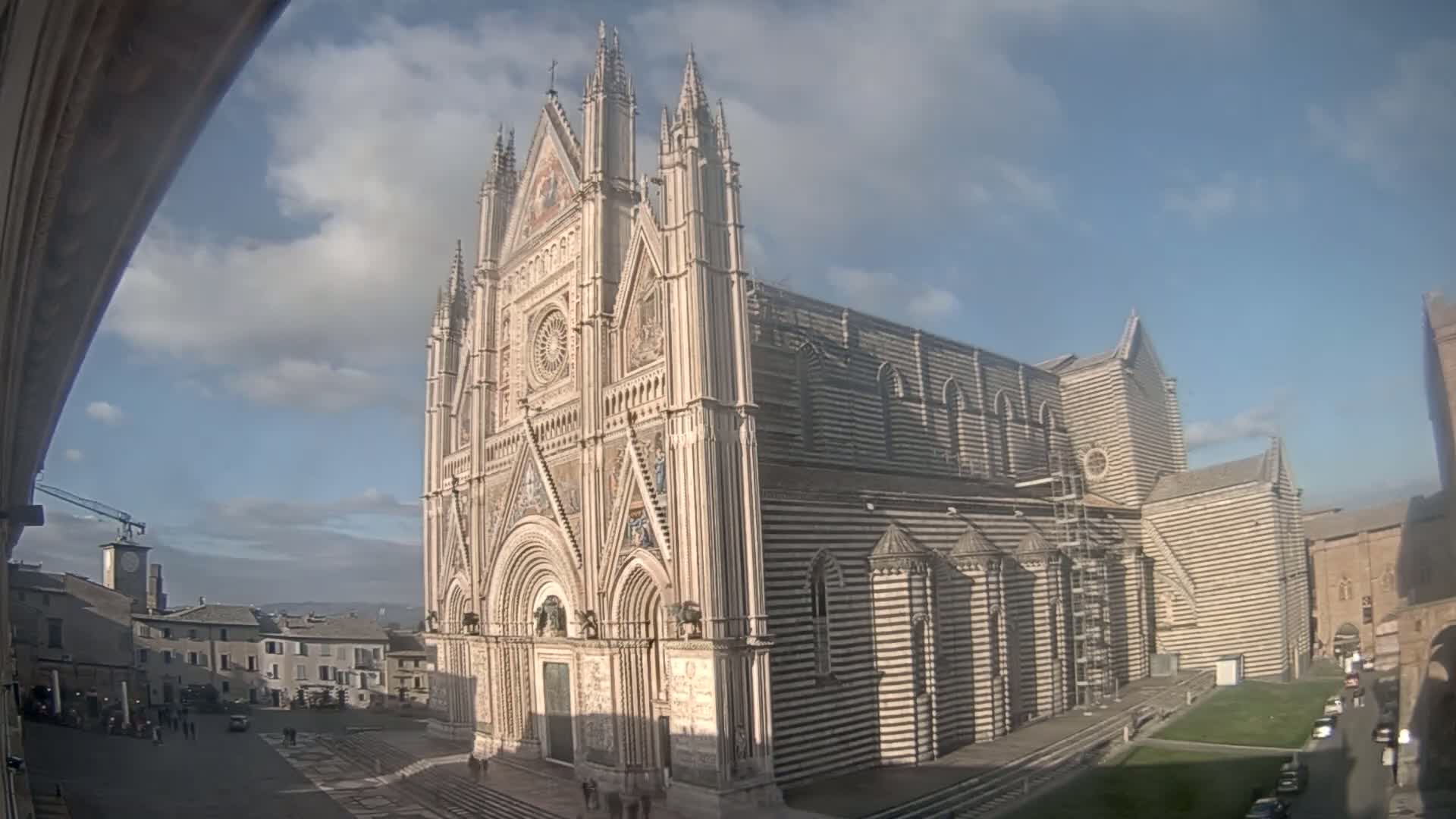 A large, striped cathedral stands in a town square under a partly cloudy sky.