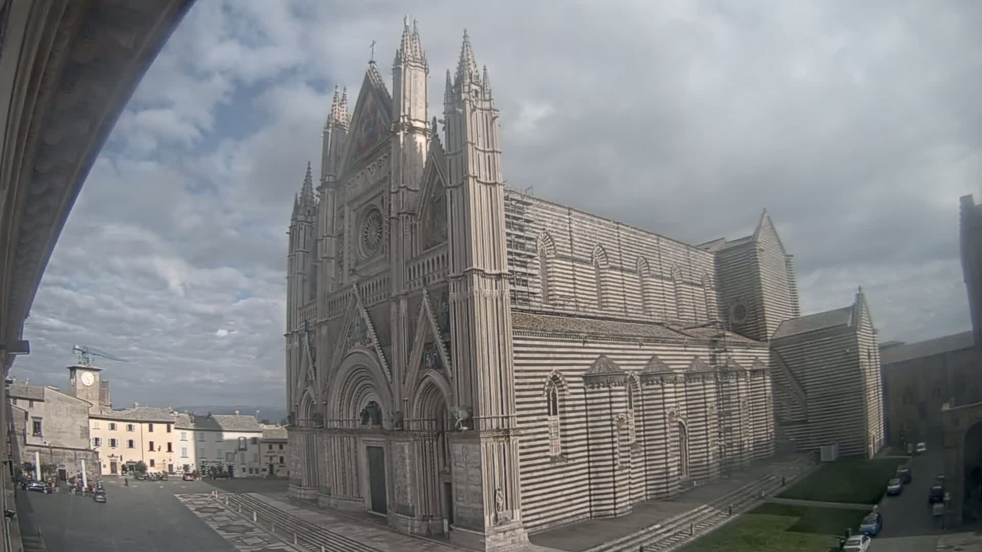 A large, striped cathedral with Gothic architecture stands in a town square under a cloudy sky.