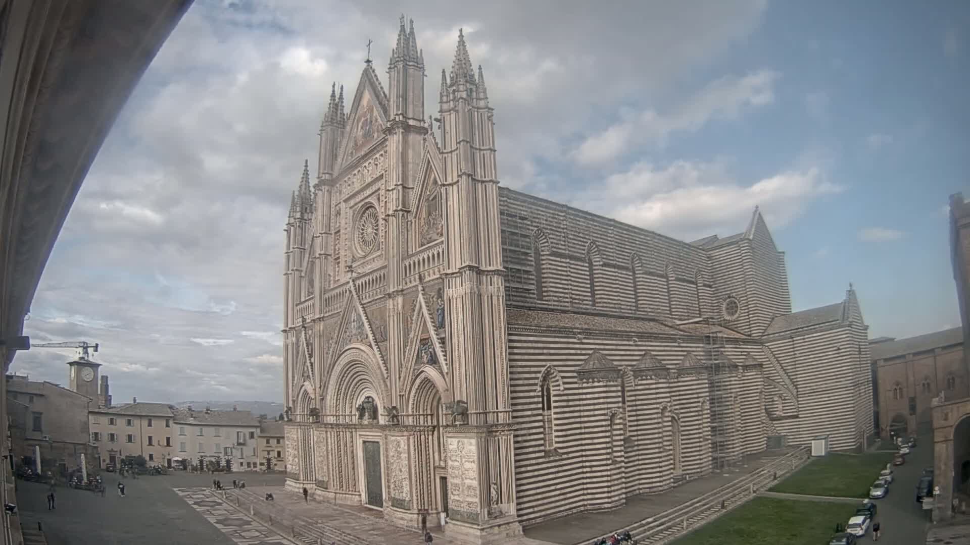 A large, striped cathedral stands in a town square under a partly cloudy sky.