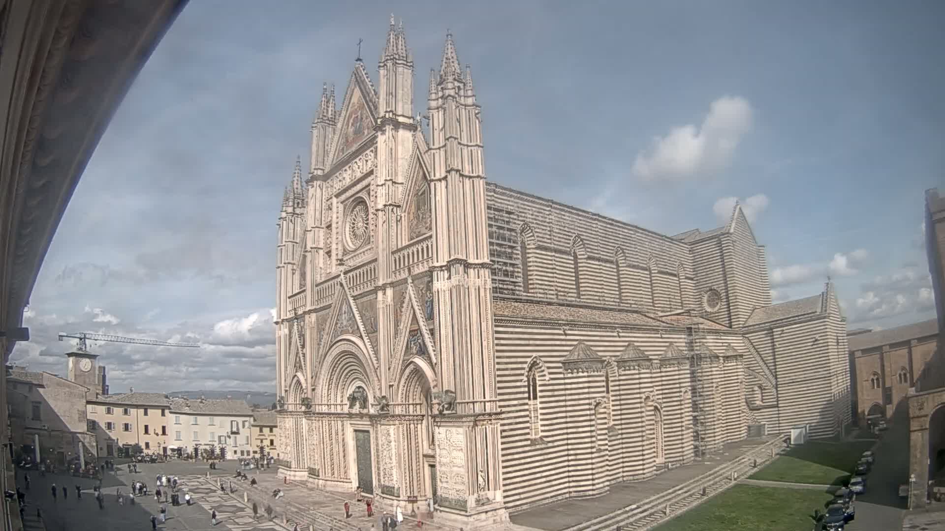 A large, striped cathedral with a Gothic facade stands in a city square under a partly cloudy sky.