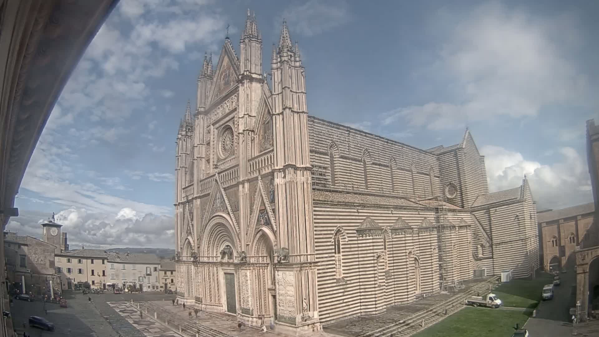 A large, striped cathedral stands in a town square under a partly cloudy sky.