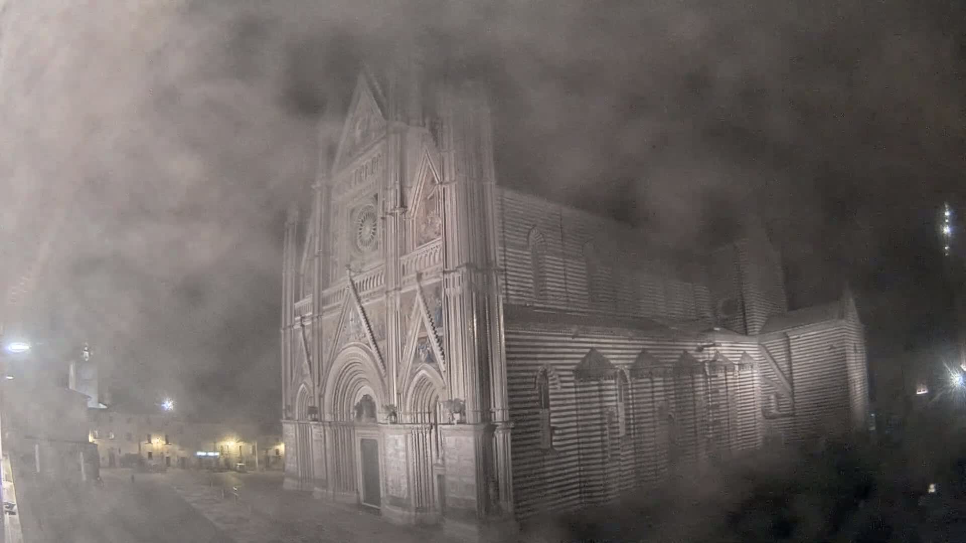 A large, striped stone cathedral stands in a town square at night, shrouded in thick fog.